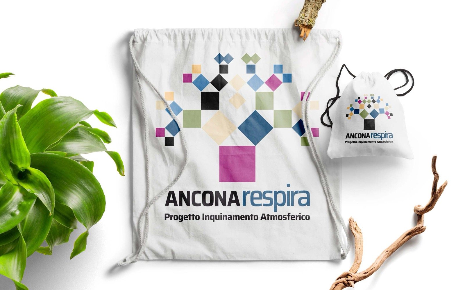 Due sacche di cotone bianco su fondo chiaro, stampate con il marchio Ancona Respira: un albero fatto di quadrati colorati, e sotto la scritta Progetto Inquinamento Atmosferico. Accanto una pianta verde e due rami secchi.