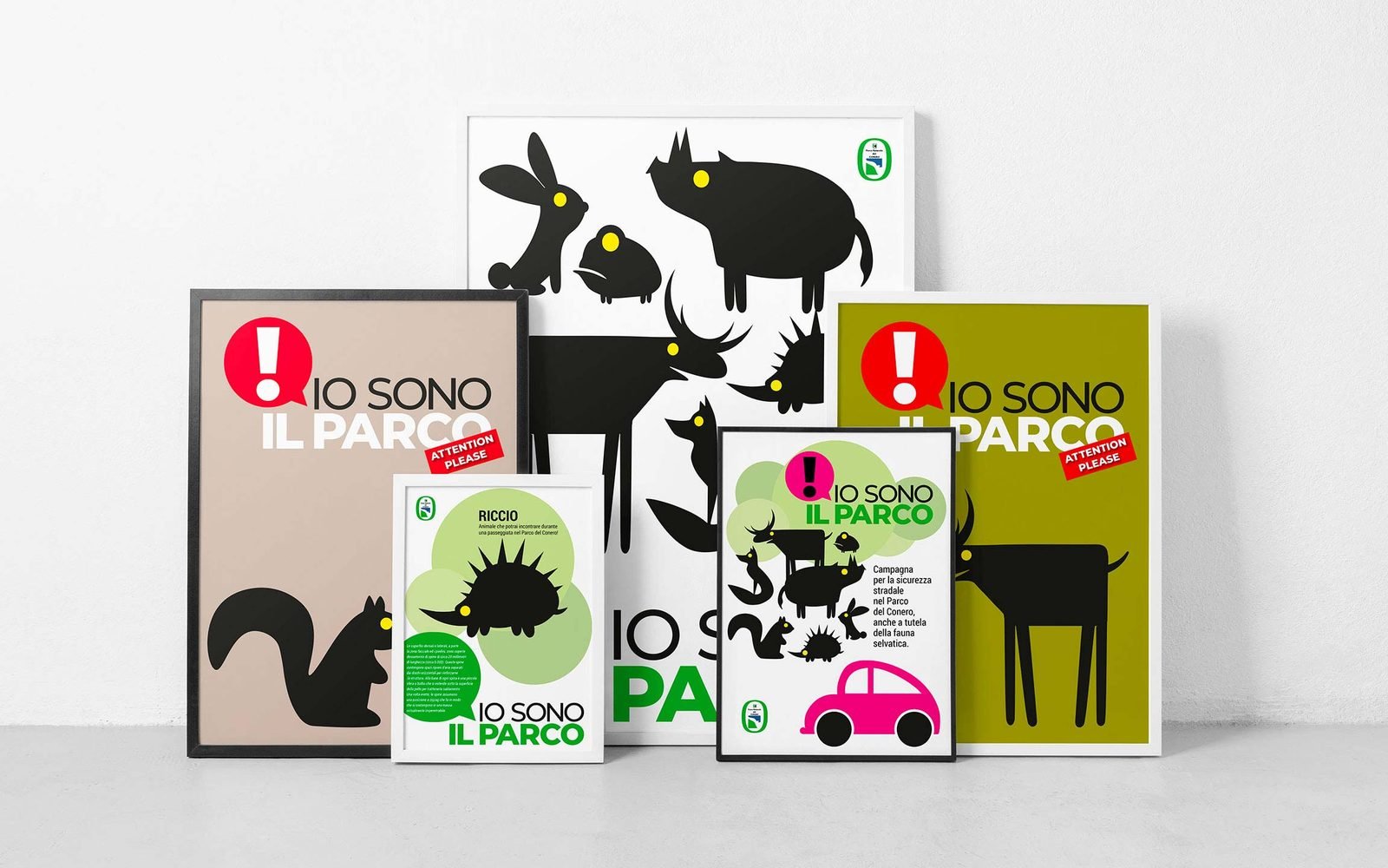 Cinque manifesti della campagna appoggiati a una parete chiara, alcuni incorniciati: quello grande bianco con gli animali in sagoma nera dall'occhio giallo, due col titolo IO SONO IL PARCO e il punto esclamativo dentro una nuvola rossa su fondo tortora e verde oliva, uno piccolo con la scheda del riccio e uno con l'auto rosa della campagna sulla sicurezza stradale.