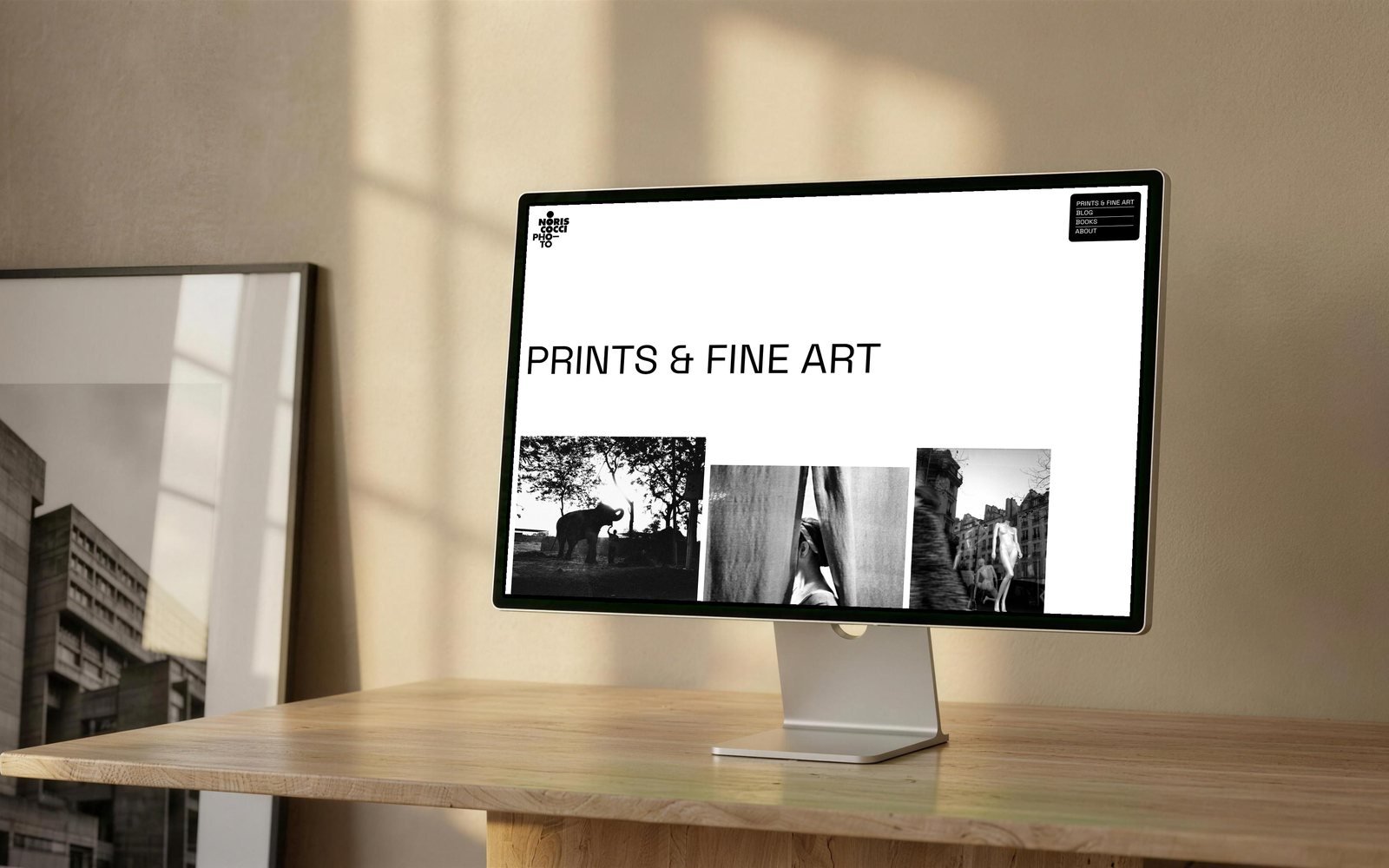 Mock-up: la sezione Prints & Fine Art di noriscocci.com su un monitor appoggiato a una scrivania di legno chiaro, con il titolo grande a sinistra e sotto tre fotografie in bianco e nero a quote diverse; a lato una stampa incorniciata appoggiata al muro.