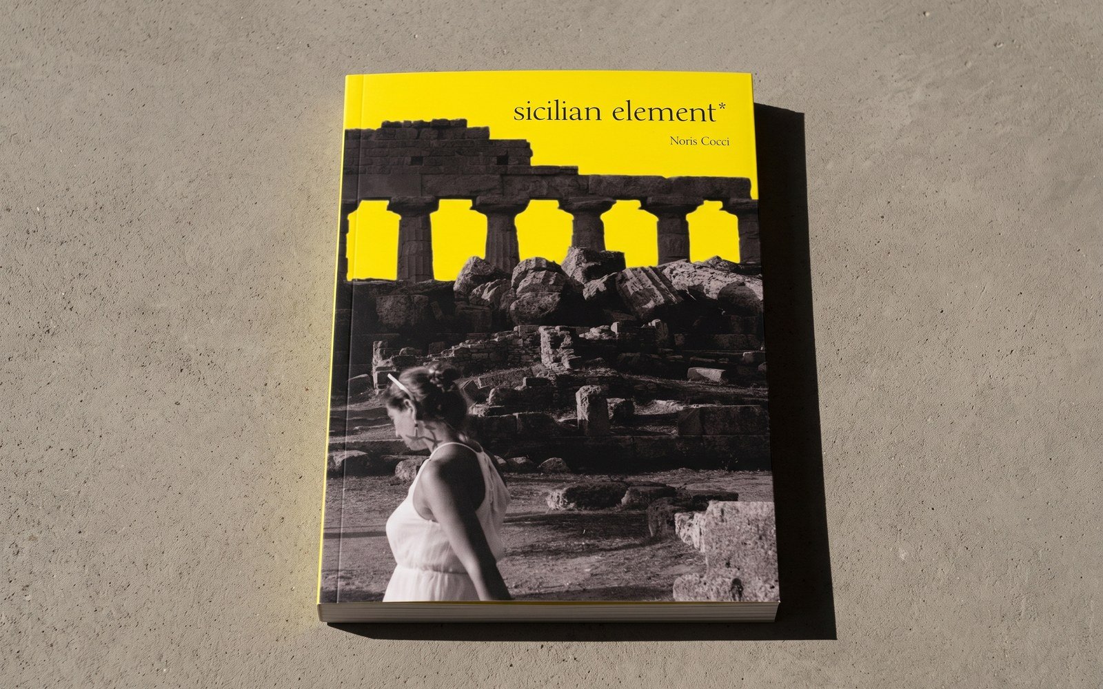 Mock-up del libro chiuso, appoggiato su un piano di cemento chiaro e ripreso da poco sopra: copertina gialla, fotografia in bianco e nero di un tempio greco e di una ragazza vestita di bianco, titolo in minuscolo sul giallo.