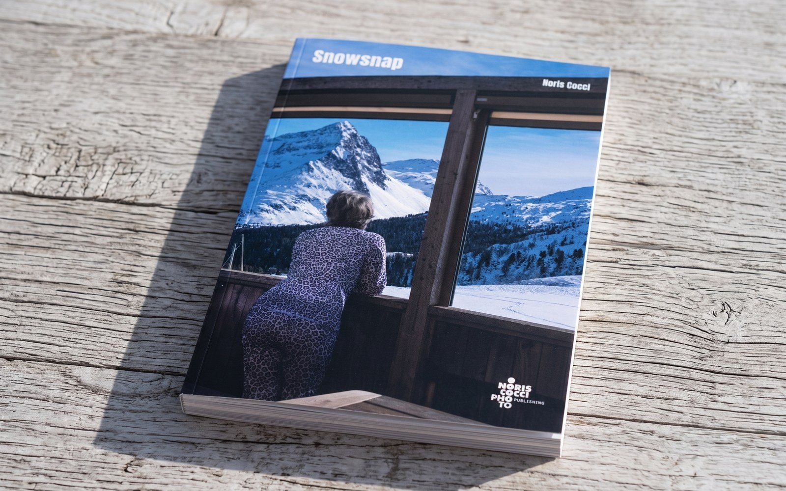 Mock-up del libro chiuso su un tavolo di legno chiaro, ripreso da poco sopra: in copertina la veduta presa da dentro un rifugio, con le vetrate che inquadrano le cime innevate, e il titolo Snowsnap in bianco.